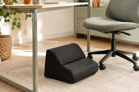 Repose-pieds de bureau noir en forme de coin placé devant une chaise, dans un espace de travail lumineux.